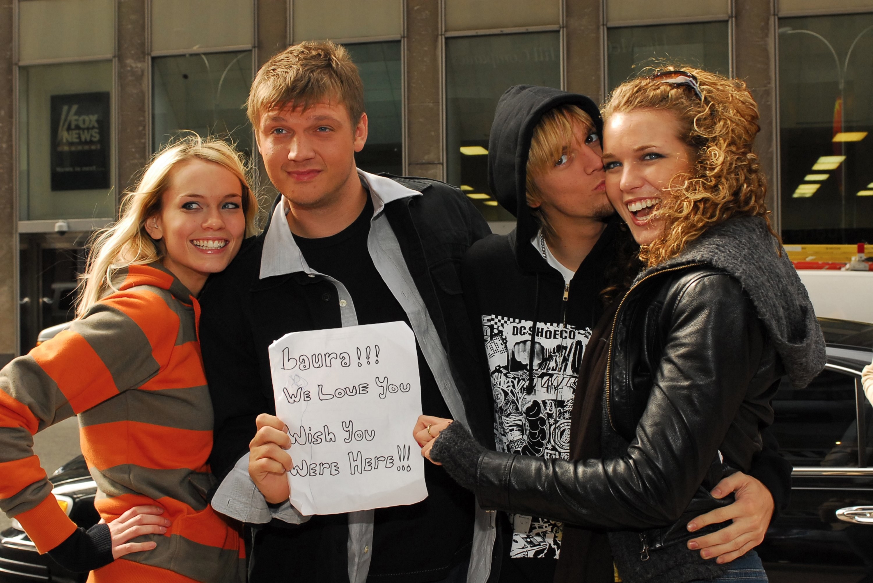 The brothers Nick and Aaron Carter stand between two smiling women, holding up a small paper sign that reads in handwritten marker &quot;laura! we love you. wish you were here.&quot;