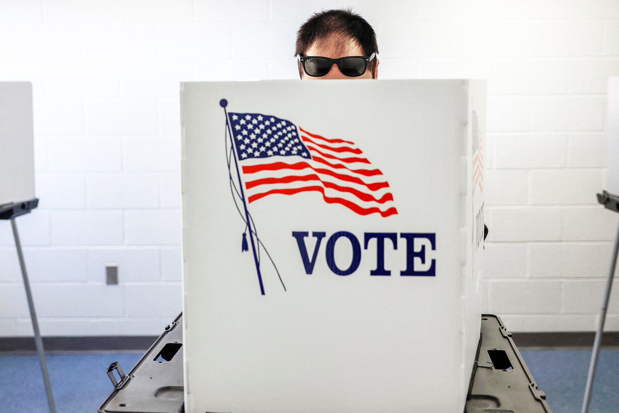 Someone with short hair and sunglasses peers over a voting booth with an American flag and the word &quot;vote&quot; on it