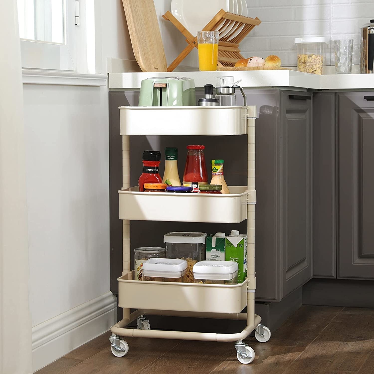 The 3-tier metal utility cart in a kitchen, holding things like a toaster, bottles of sauce, containers of food, and other kitchen items.