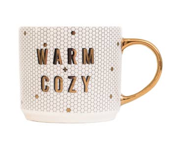 a white tiled mug that says warm + cozy on it in gold