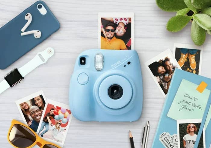 Blue mini camera printing a photo, surrounding by photos, scrapbook, phone, sunglasses and watch