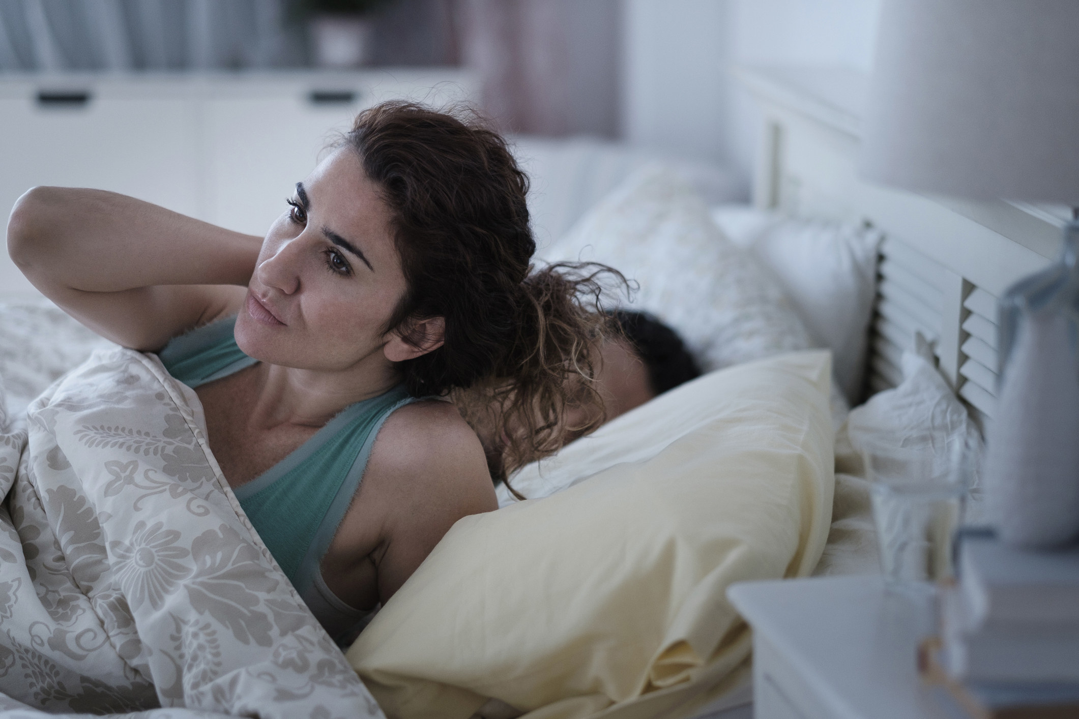 a woman looking distressed and sitting up in bed as a man sleeps next to her