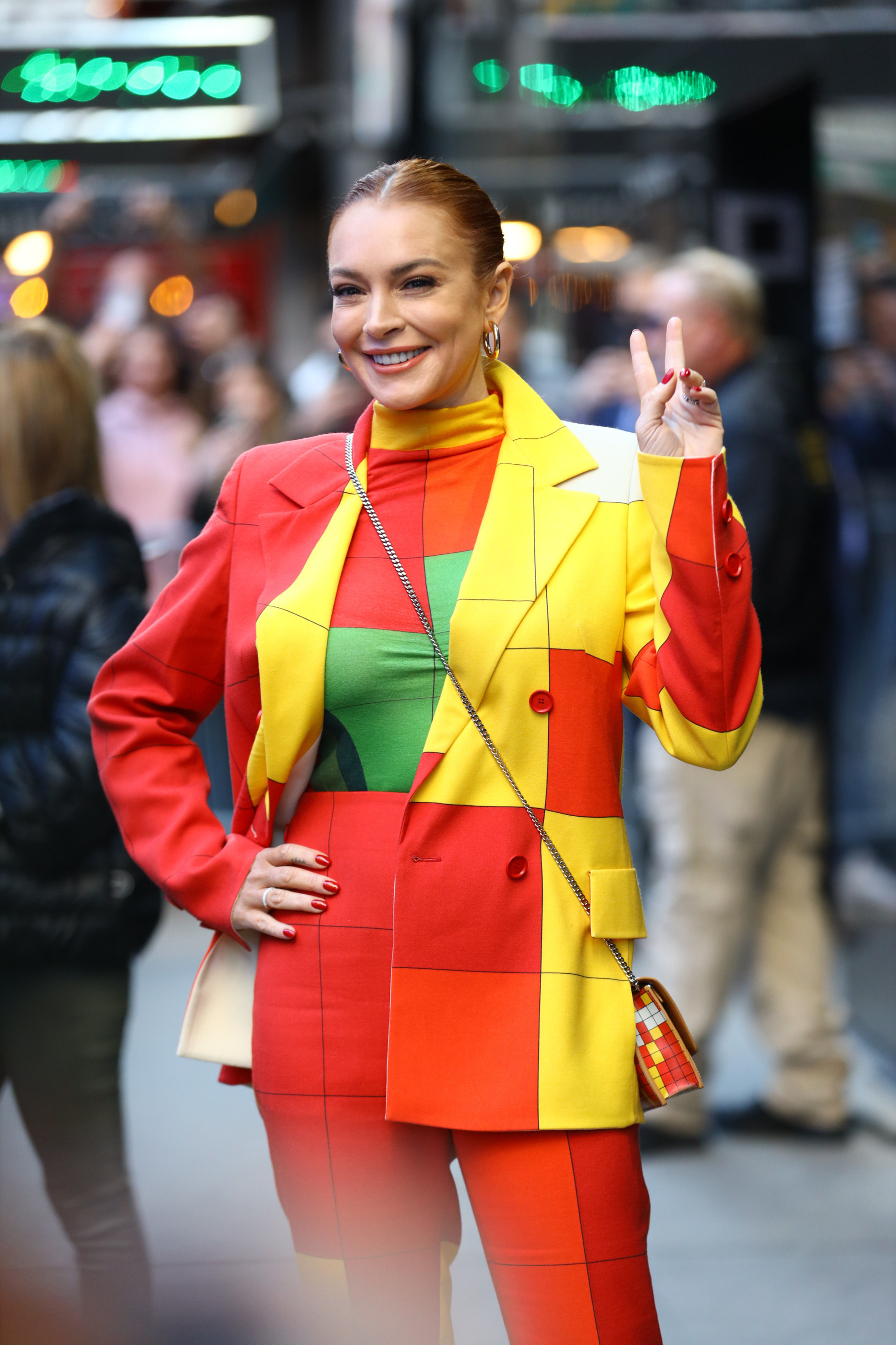 Lindsay smiling and giving the peace sign. She&#x27;s wearing a color-blocked pantsuit and her hair pulled back into a sleek bun