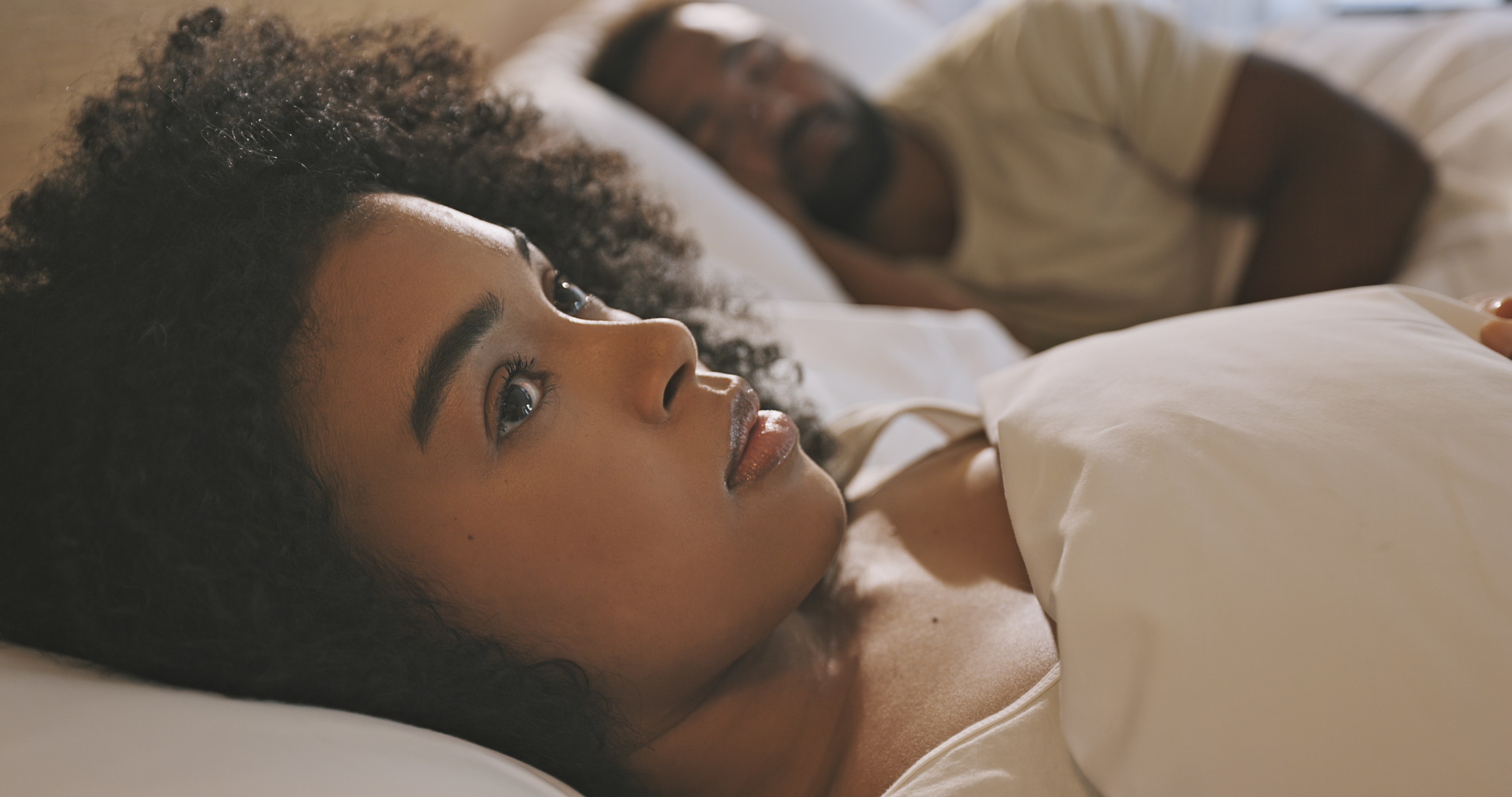 a woman lying awake in bed next to her sleeping partner