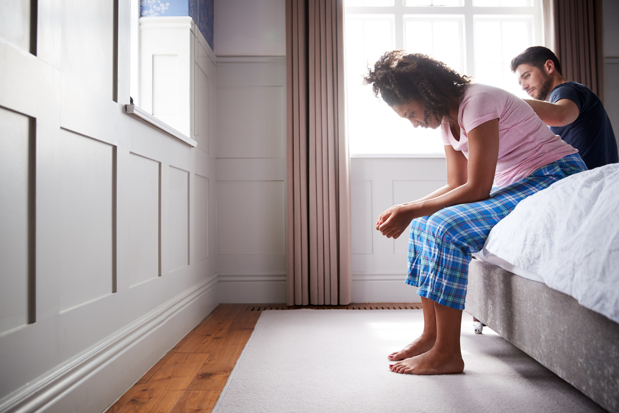 a woman sitting on the edge of a bed looking distressed as her partner rubs her back