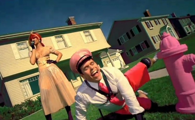 Perez Hilton is wearing a suit and a leather harness, cocking his leg and peeing on a pink hydrant outside a suburban house, Rihanna stands behind him in a gown