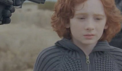 A redhead child looks scared as they stand in a desert with a gun pointed at their head