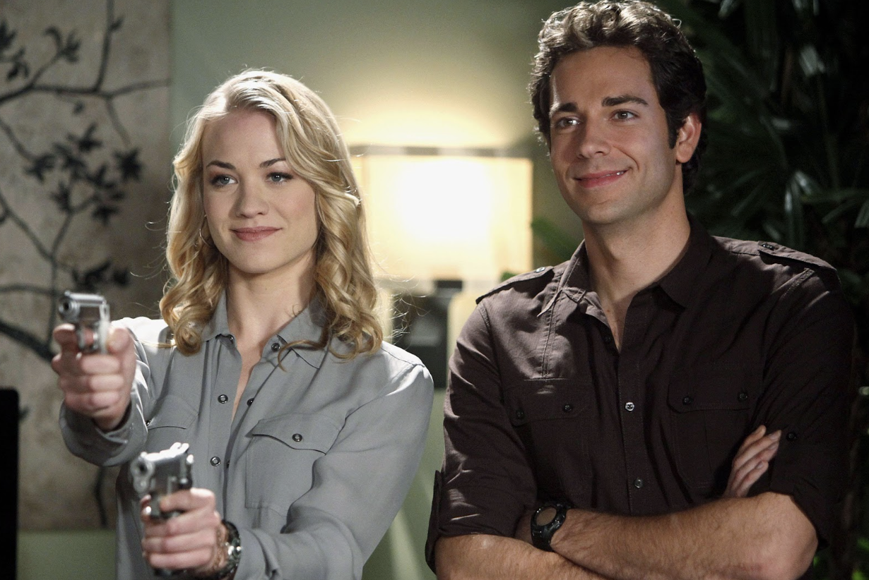 A blonde woman holds two guns while standing next to a grinning man