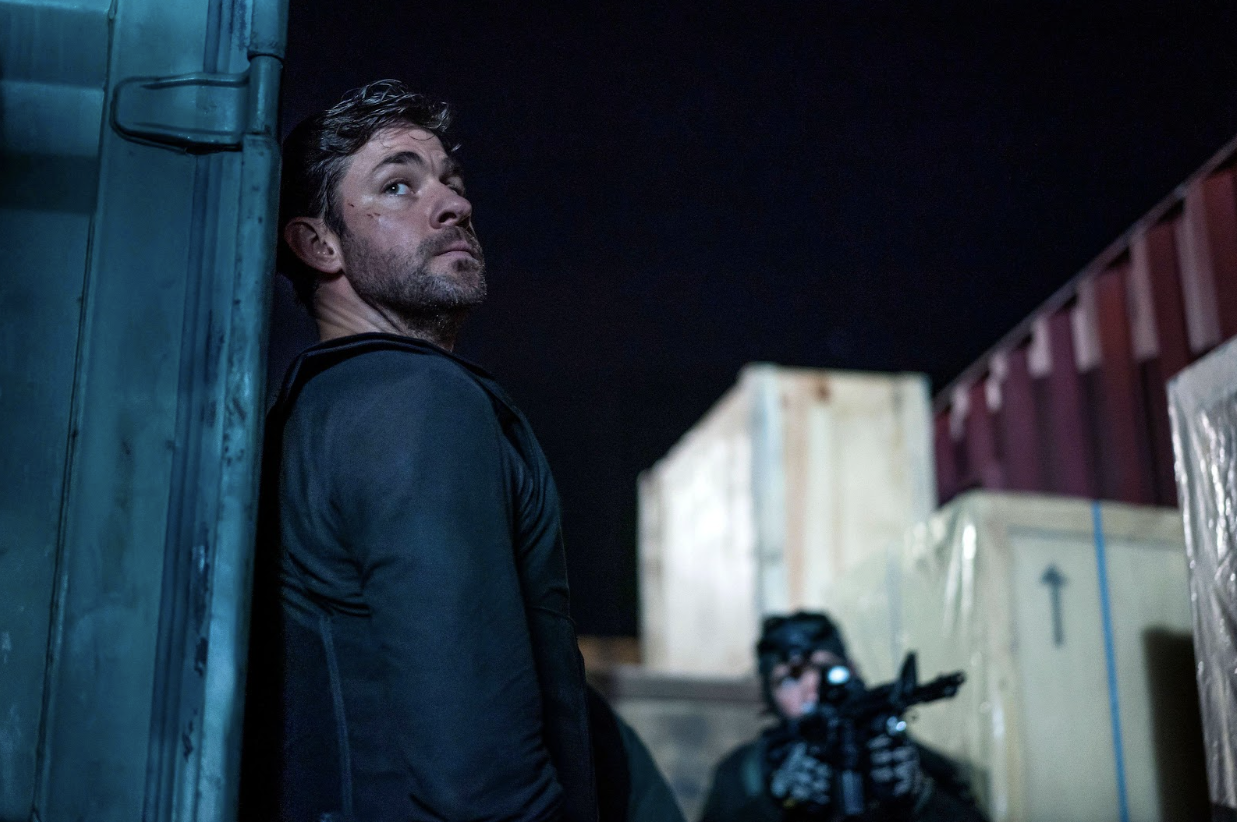 A man with scruffy facial hair and a body armor vest stands with his back to a shipping container near a gun-toting soldier