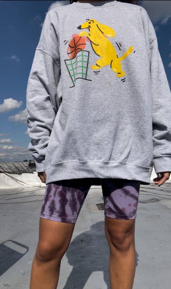 a model on a rooftop wearing the grey sweatshirt with art of a yellow dog dunking a basketball in a green trash can, and purple tie-dye bike shorts