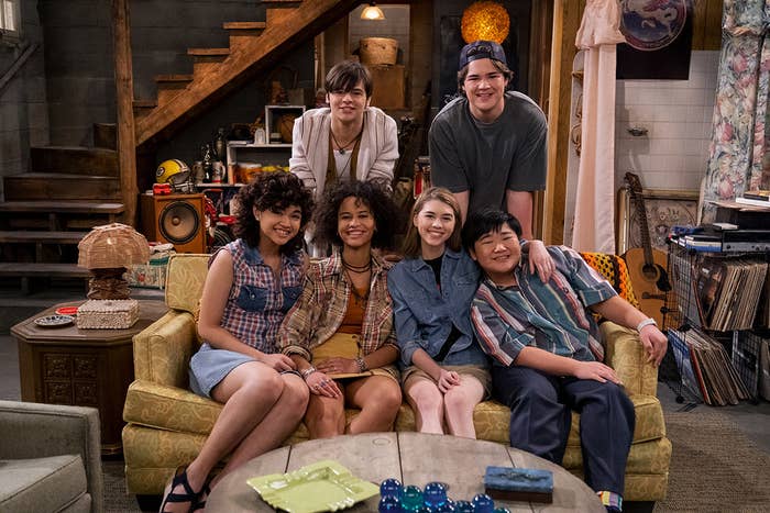 The gang of That 90s Show smiles and sits on the couch in the basement