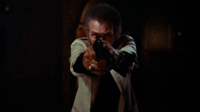A man in a white overcoat and hand jewelry points a gun at the camera
