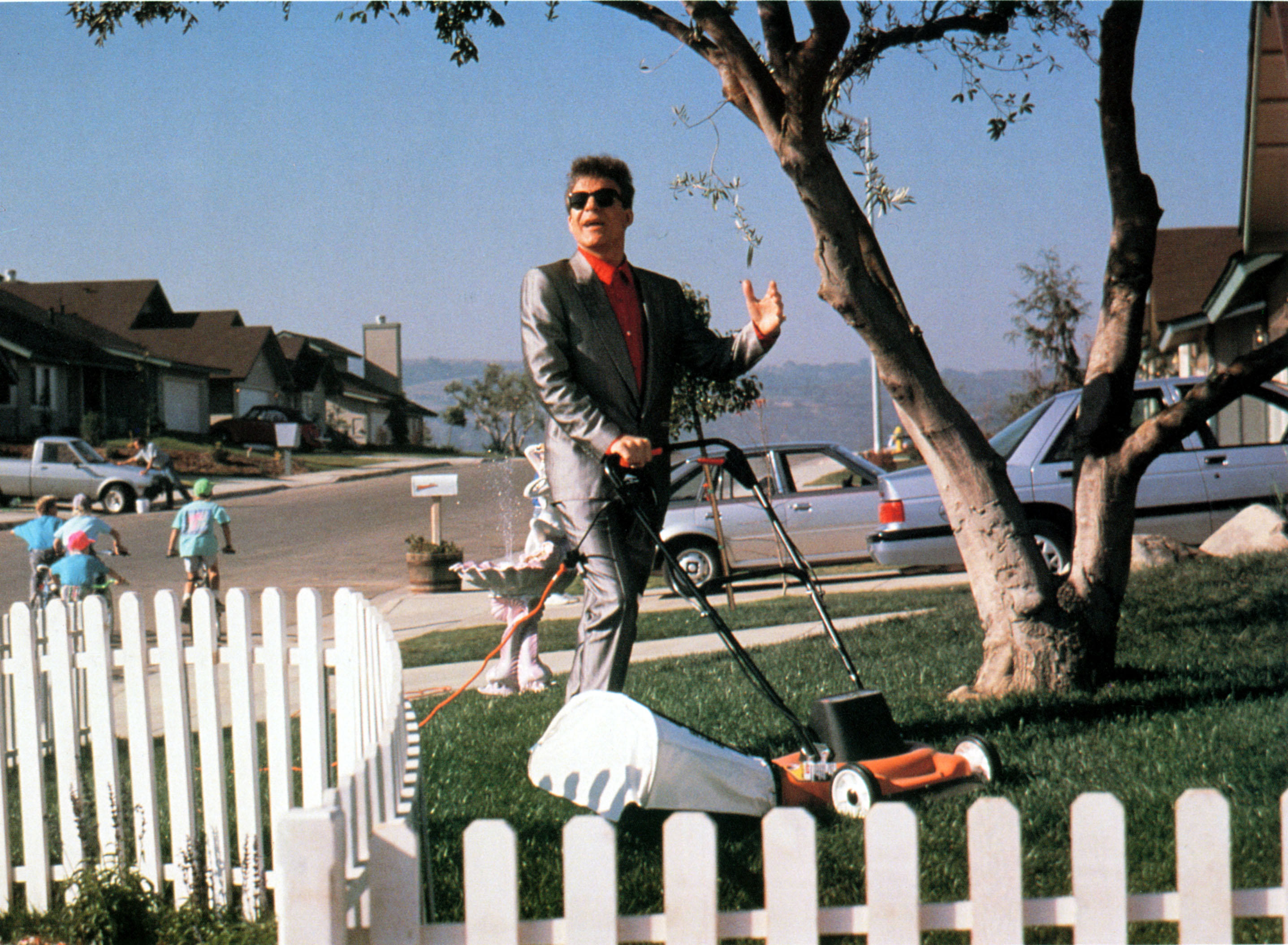 A gangster in a colorful three-piece suit mows his lawn in a suburban neighborhood
