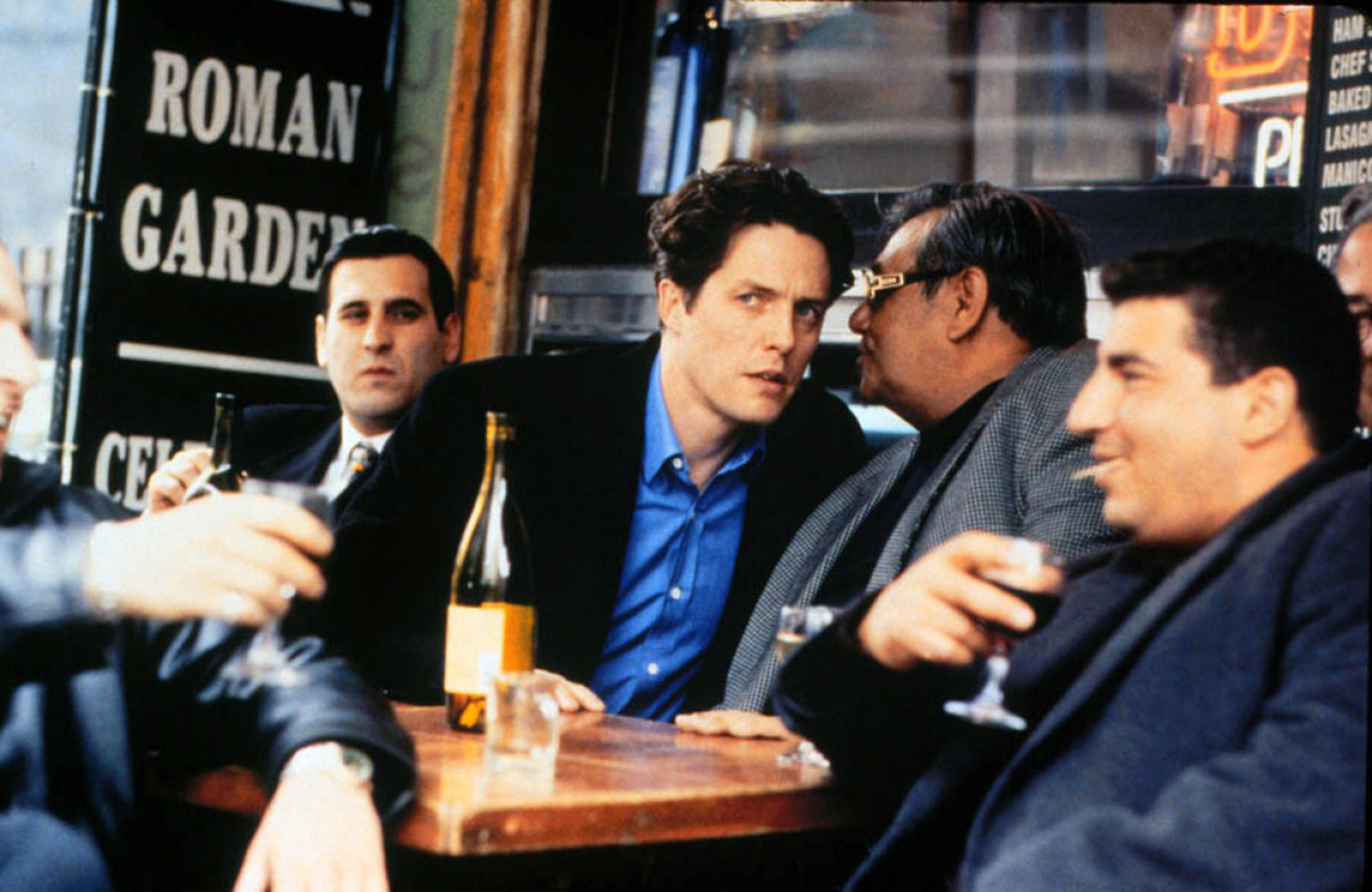 A confused young man in a black jacket and blue shirt stares out while having a drink with well-dressed gangsters