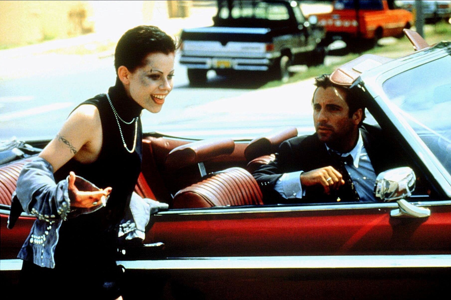 A short haired woman with messy make-up wears a wicked grin while a confused man in a convertible looks on