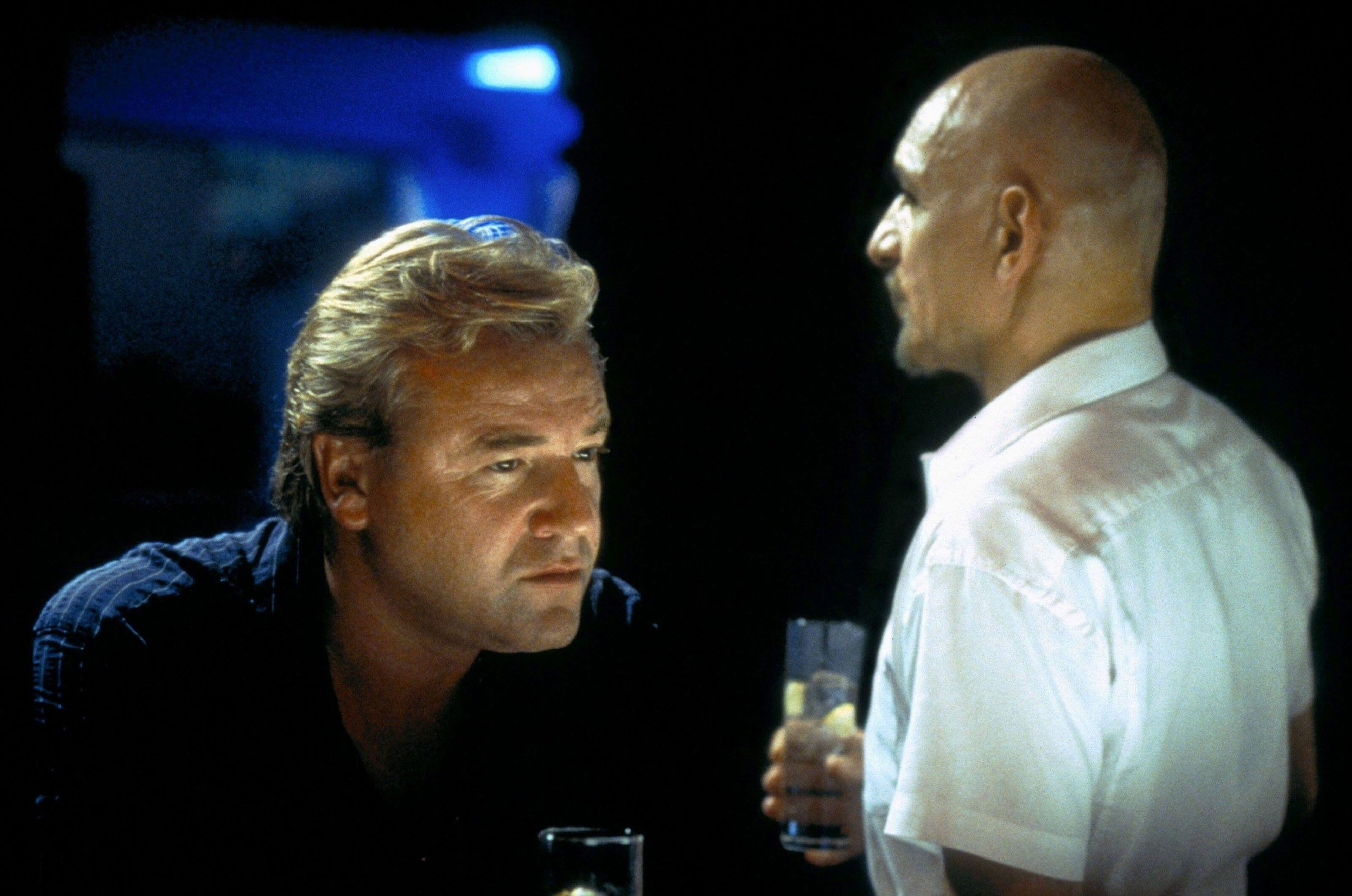 A blonde man in a dark shirt shares a drink with a bald man in a white shirt