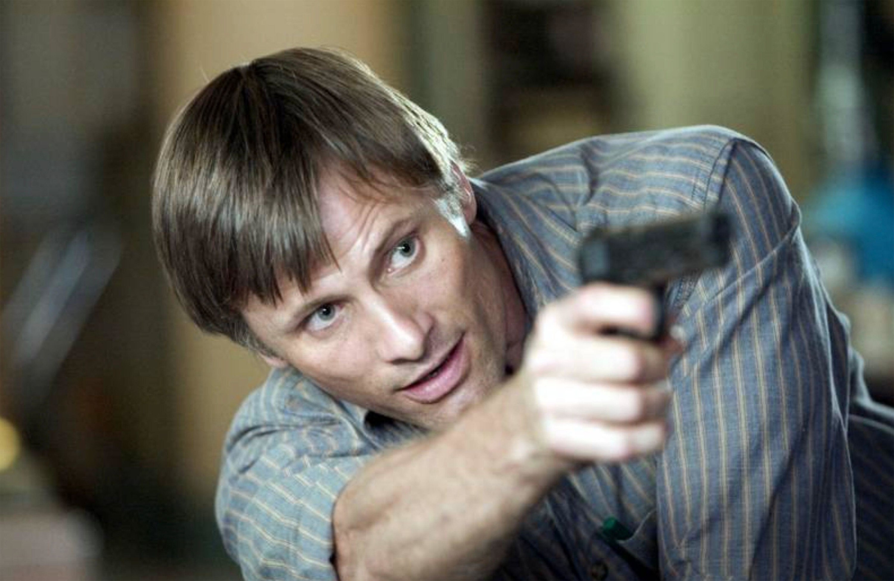 A man in a striped, button-up shirt begrudgingly points a gun at a threat