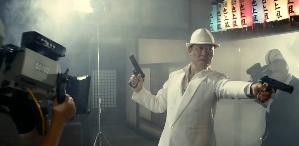 A gangster in a white suit duel wielding pistols poses for a pair of cameras
