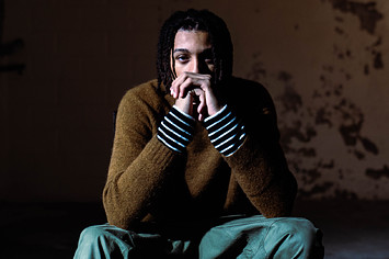 Toronto artist Kofi crouching, wearing a brown sweater with a striped undershirt and green pants.