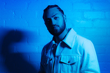 A blue-tinted photo of Ali Gatie in a jean jacket, standing against a brick wall