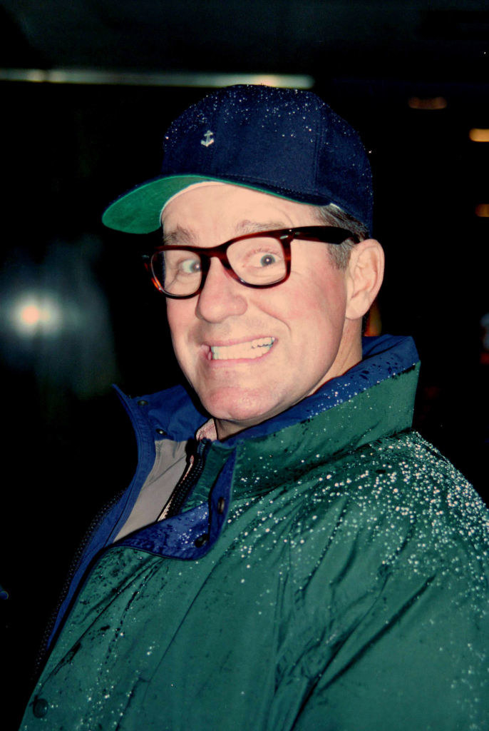Phil Hartman wearing a hat and glasses, smiling in the rain