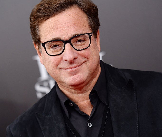 Bob Saget on a red carpet wearing glasses and smiling