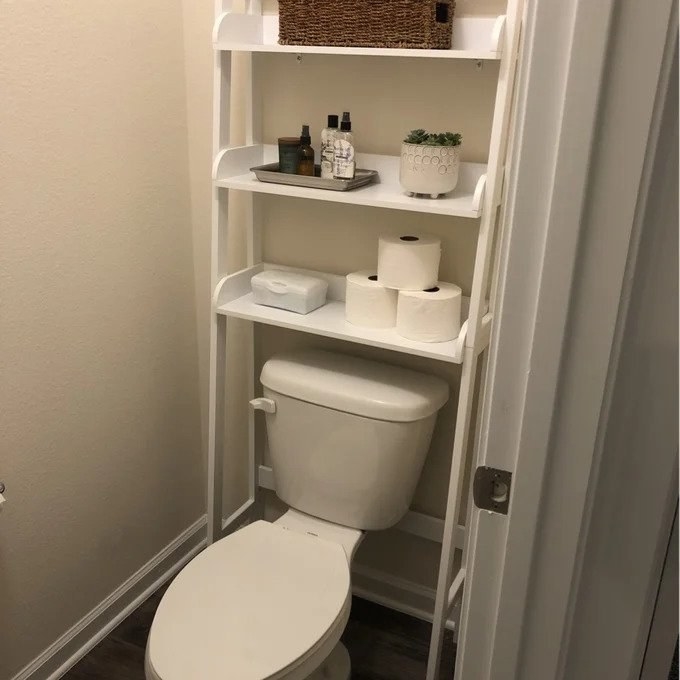 White over-the-toilet unit in bathroom with toilet paper, tray, planter, and basket on shelves of the unit