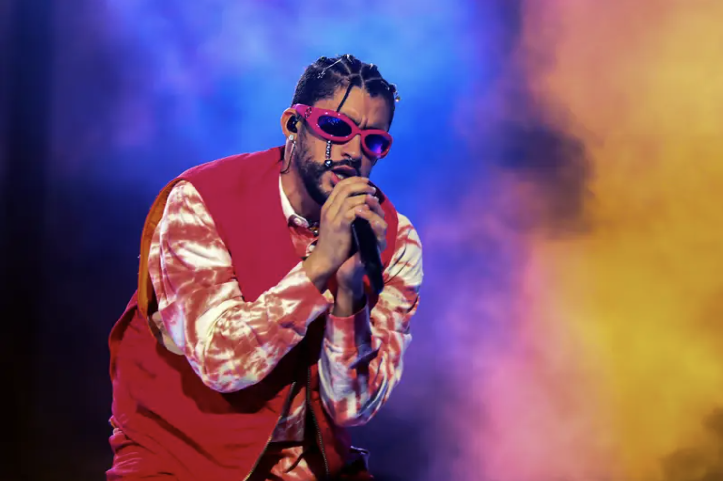 Bad Bunnie in a red vest, red tie dye long-sleeve shirt, and red sunglasses singing into a mic