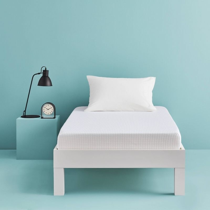 A white mattress on a white bed frame in a blue room with black lamp and clock