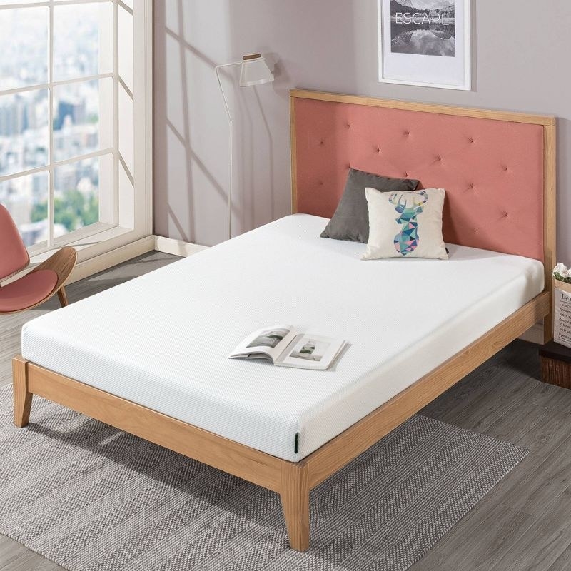 A pink and brown bed frame with a white mattress, magazine and colorful and grey pillow