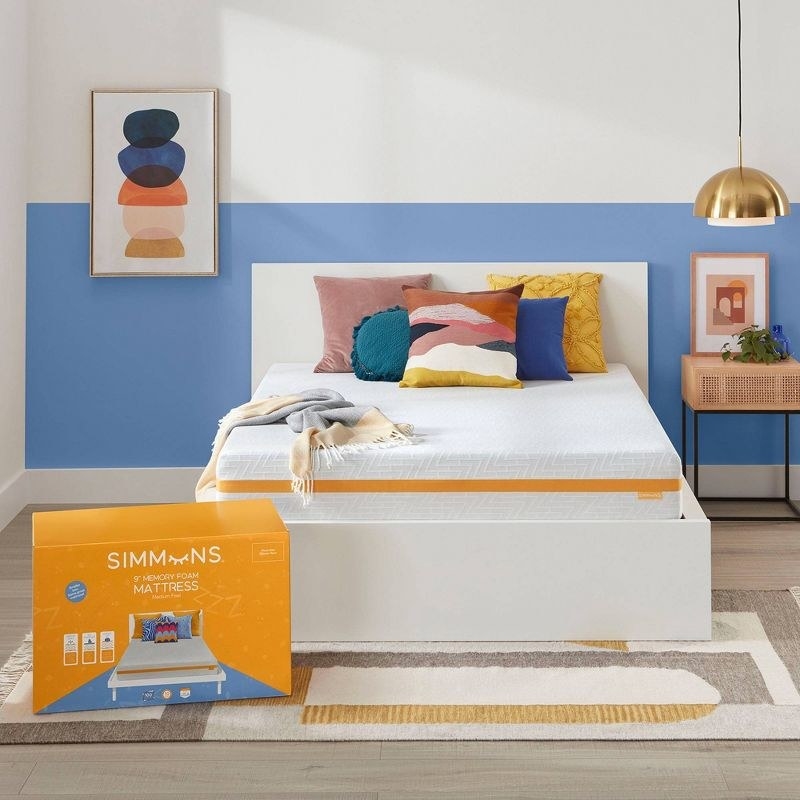 A yellow mattress box in front of a white bed with mattress with colorful blanket and pillows