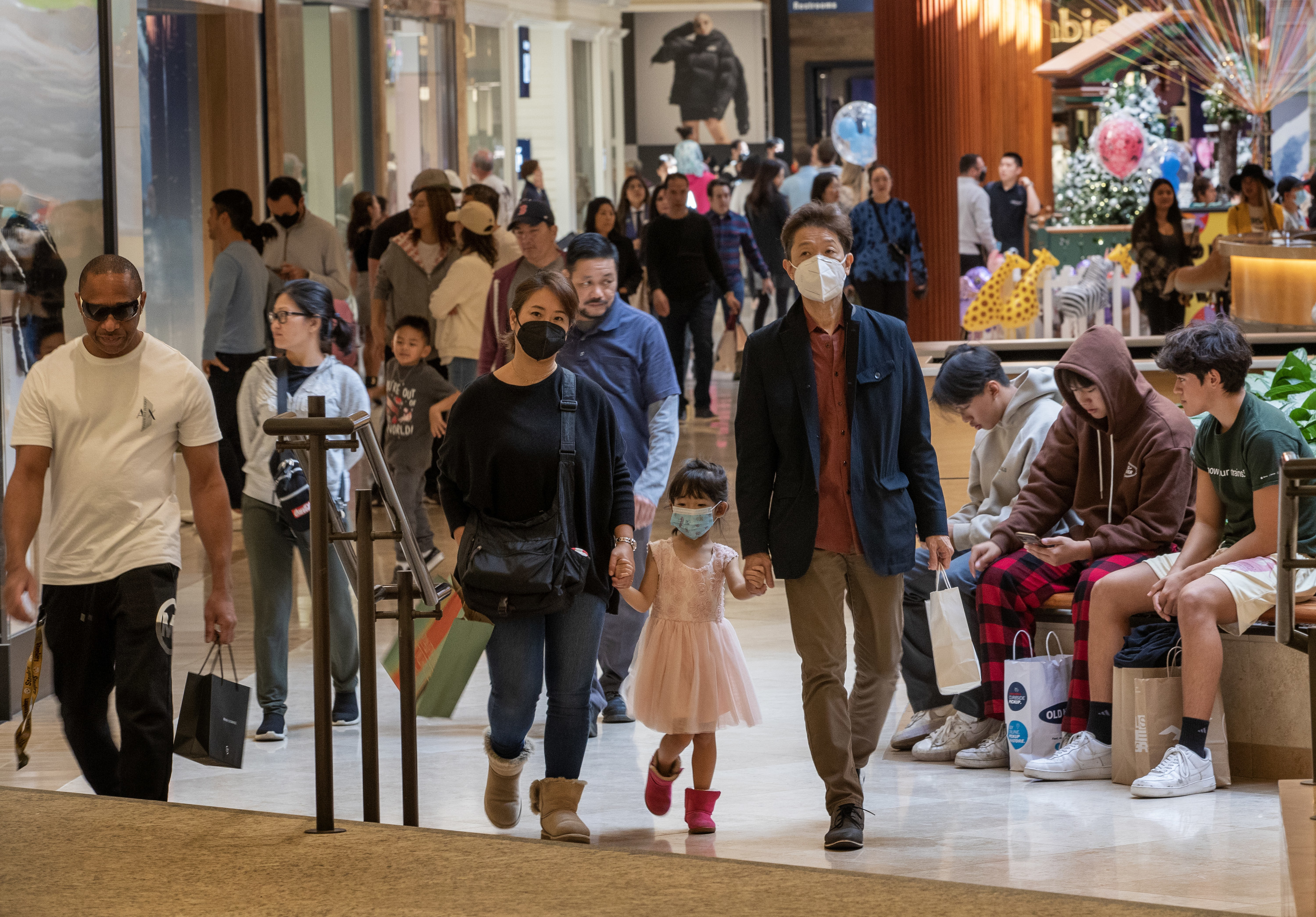 people walk through a crowded shopping mall, with only a few wearing masks
