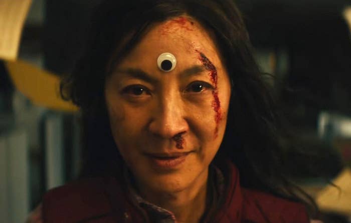 Michelle Yeoh as Evelyn looks into the camera. She has a cut on her face with blood coming out and a googly eye on the center of her forehead
