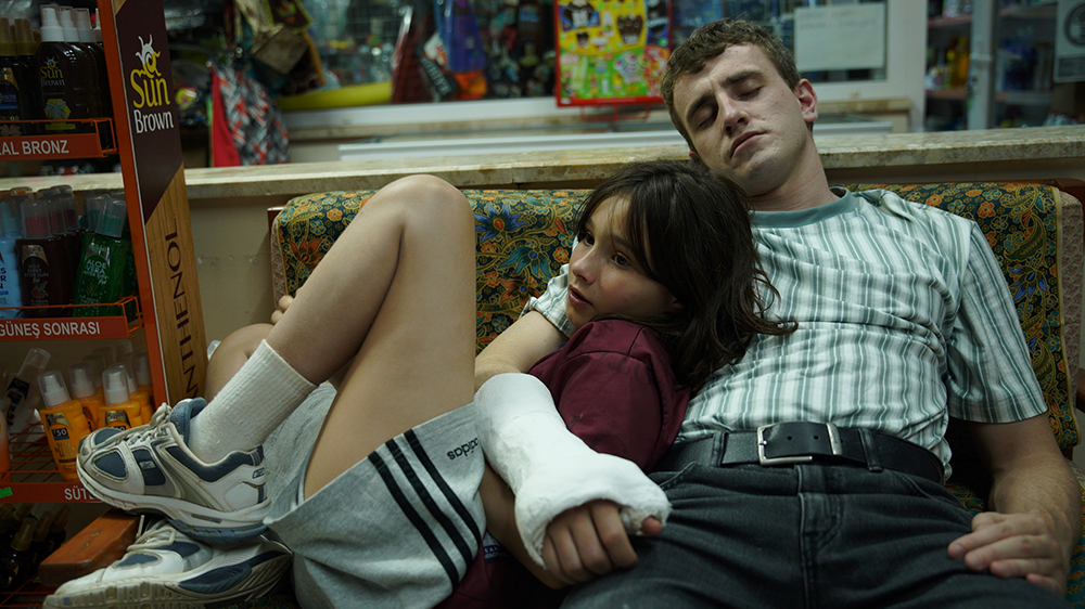 A still from Aftersun, Sophie (Frankie Corio) and Calum (Paul Mescal) sit on a couch. Calum has a cast on his arm which is slung around Sophie.