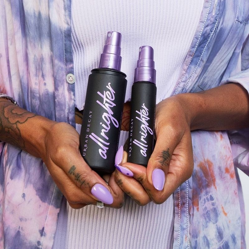 A person with a purple manicure holding small and large bottles of makeup setting spray