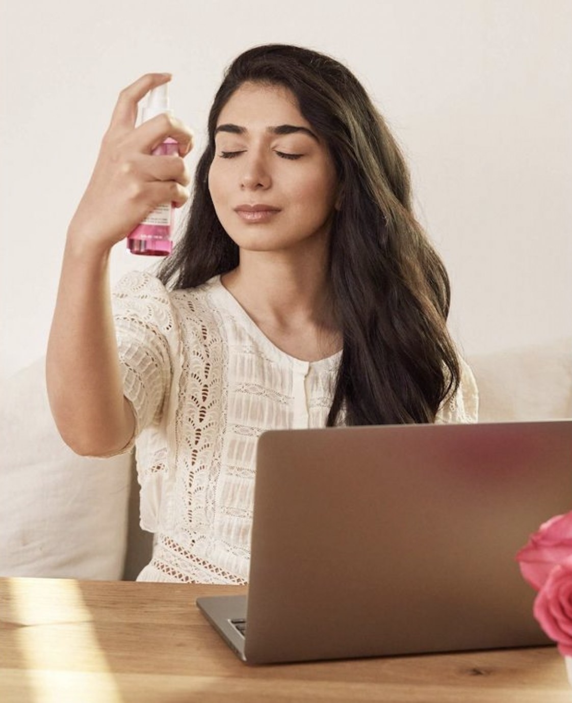 A person sitting at a table in front of a laptop spritzing facial mist on their face