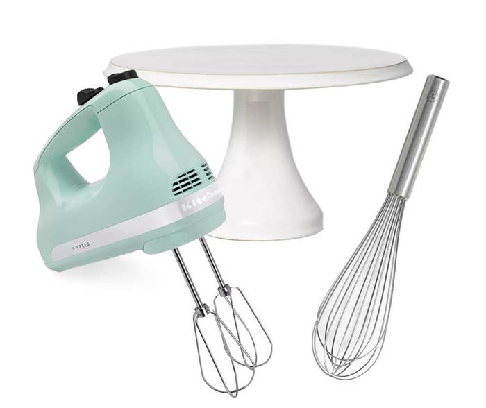 A blue KitchenAid hand mixer, a white, ceramic cake stand, and a metal hand whisk