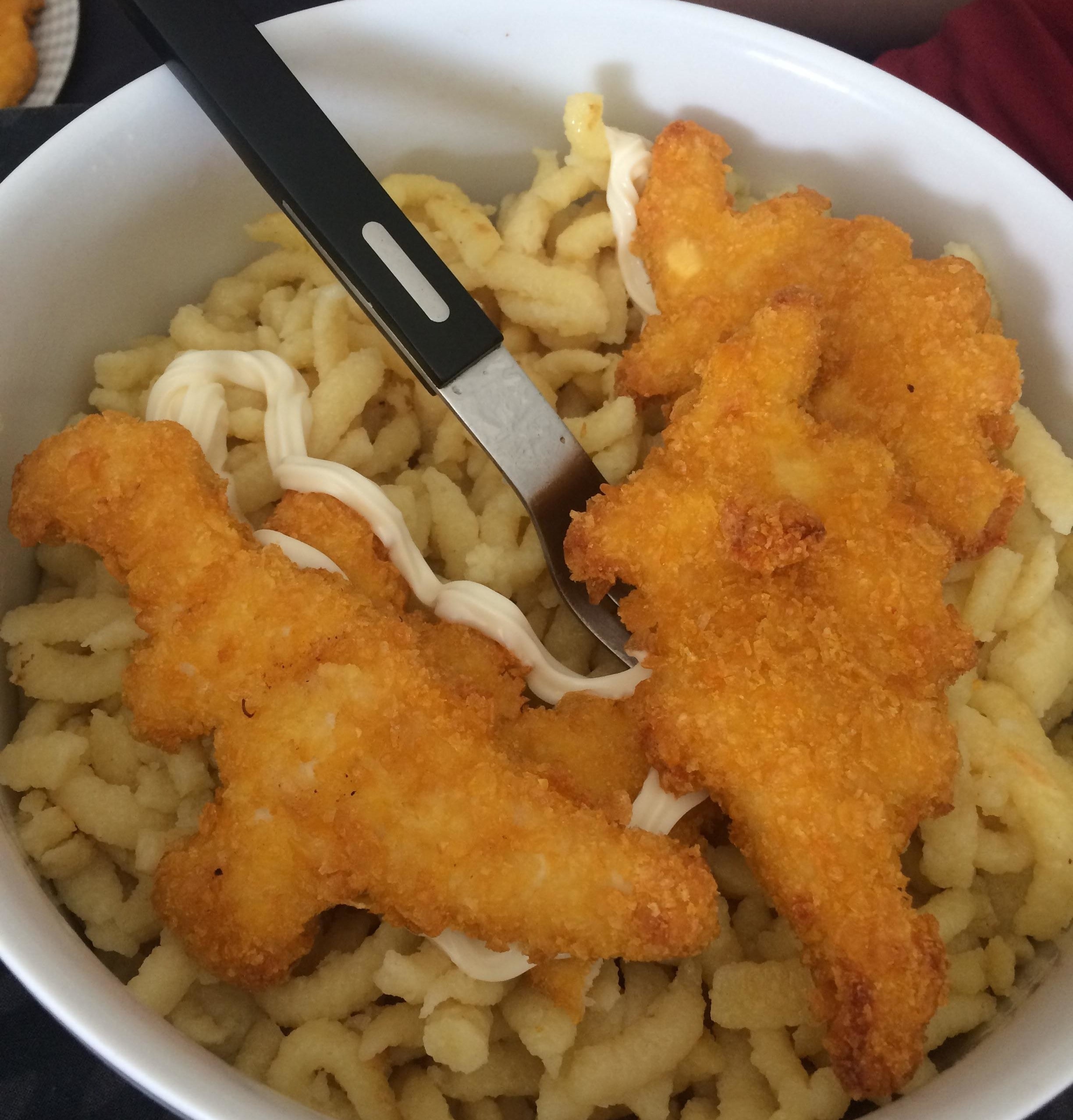 A bowl with noodles, 3 dino nuggets, and mayo drizzled on top
