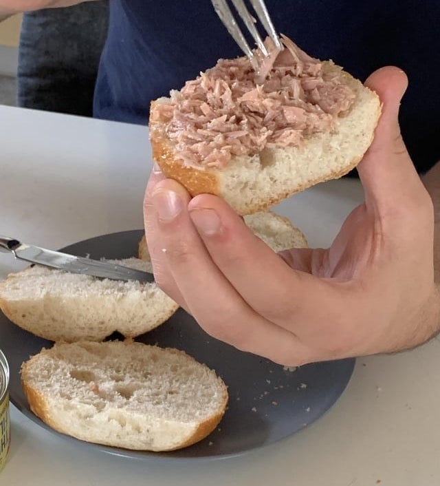 A person using a fork to spread tuna on a bread roll