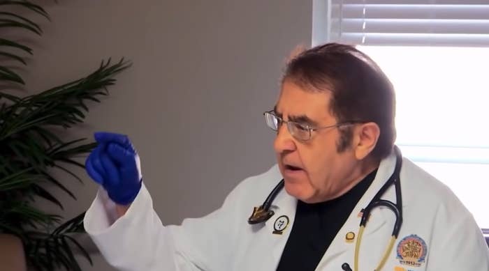 Dr. Nowzaradan wears blue latex gloves, a white doctors coat, and a strethoscope. He is scolding a patient. He is an old man with brown, wispy, dyed hair.