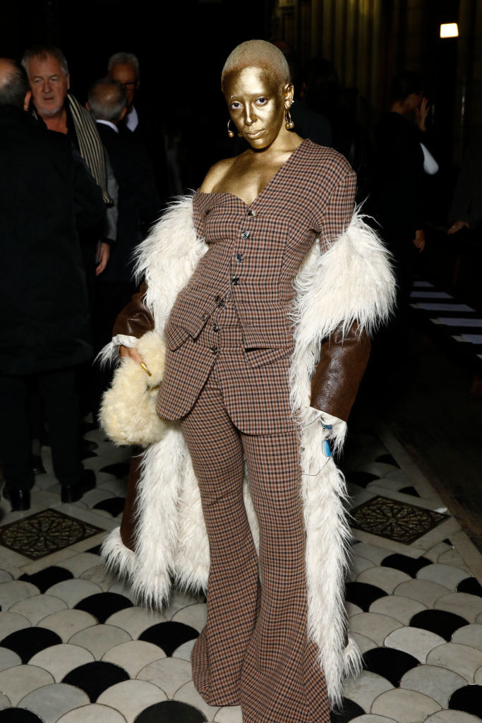 Doja in gold face and body paint and a plaid pantsuit with flared bottoms and long coat