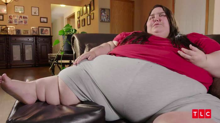 An obese woman wearing a bright pink shirt and gray pants. She sits on a dark brown sofa. She has straight brown hair.