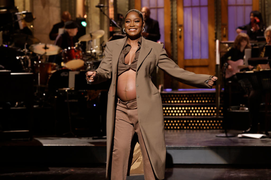 Smiling Keke in a midriff-baring tie-up top, pants, and long coat