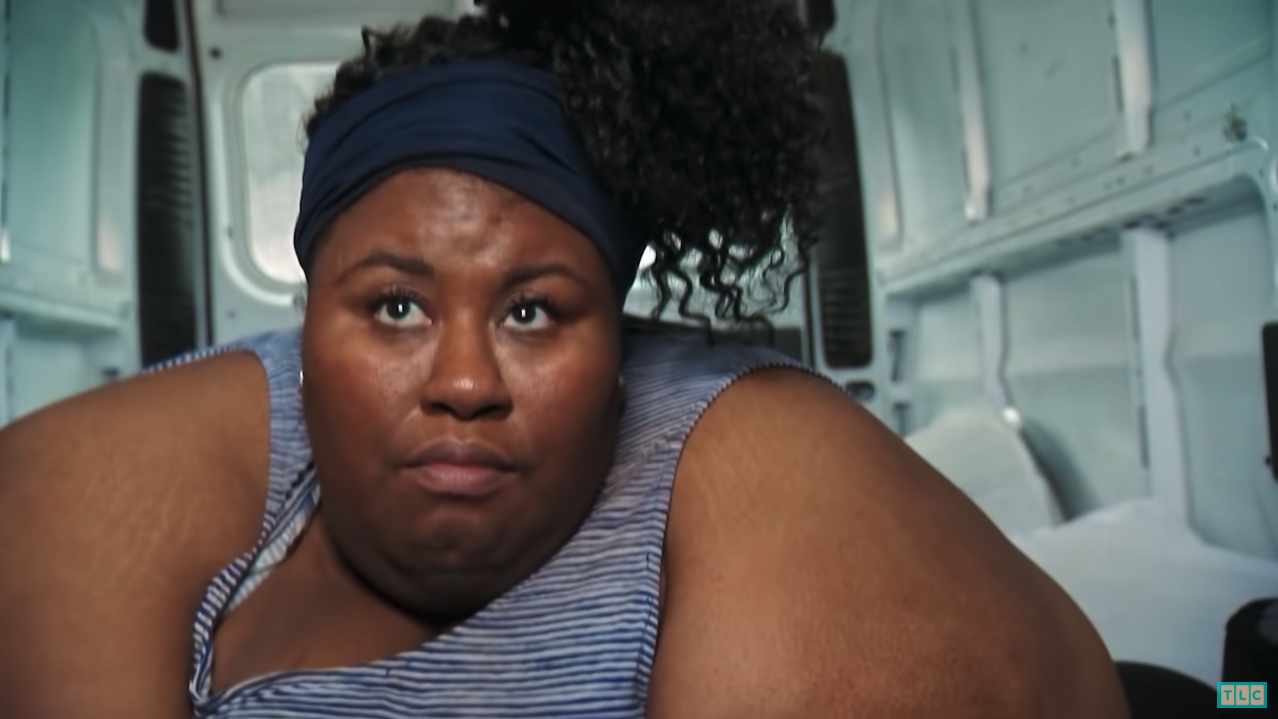 Octavia, a woman with cury black hair, a navy blue headband, and a stripped blue and white tank top, lays in the back of a moving van on a mattress. She is looking worried