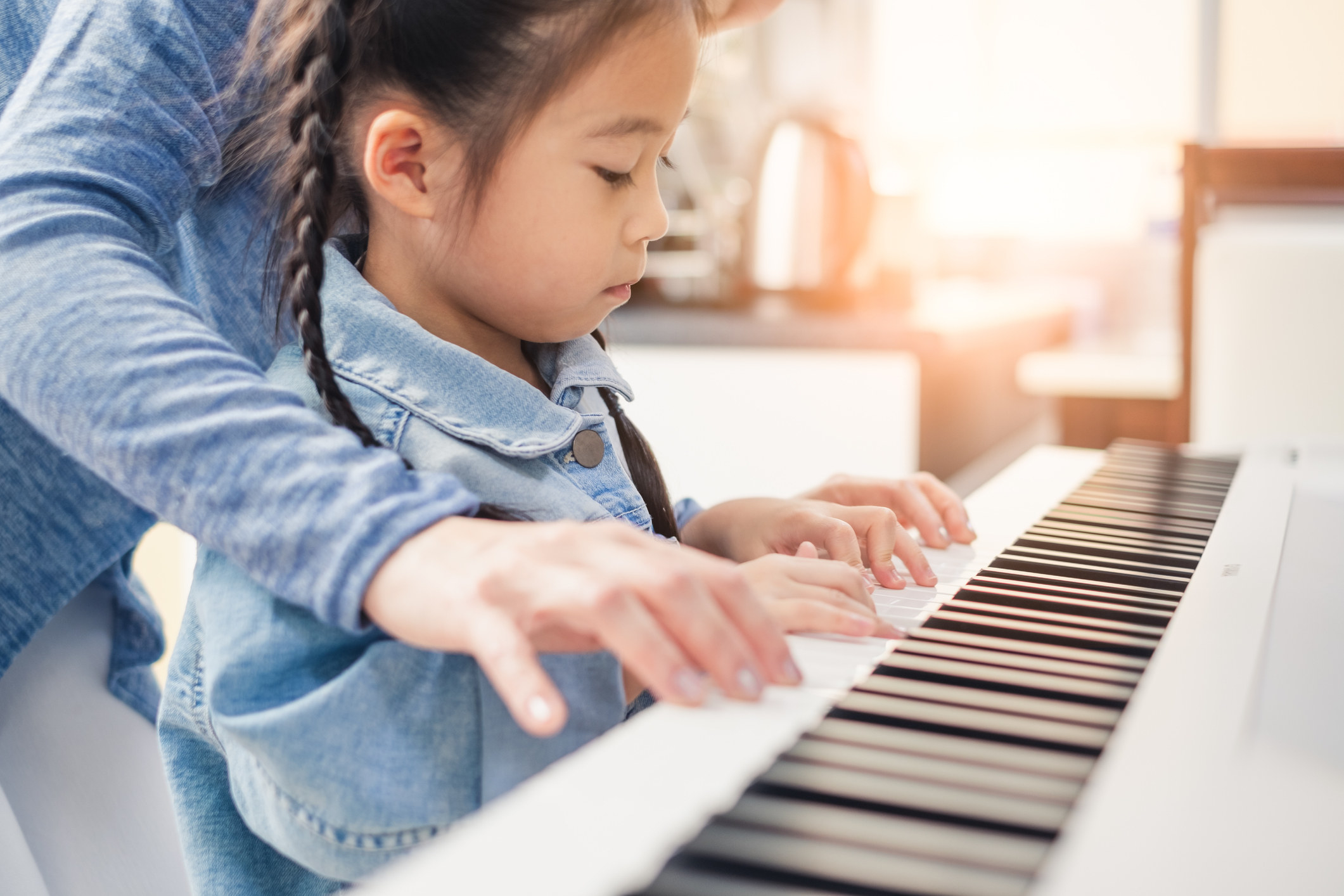 Child playing piano as adult stands over her also playing