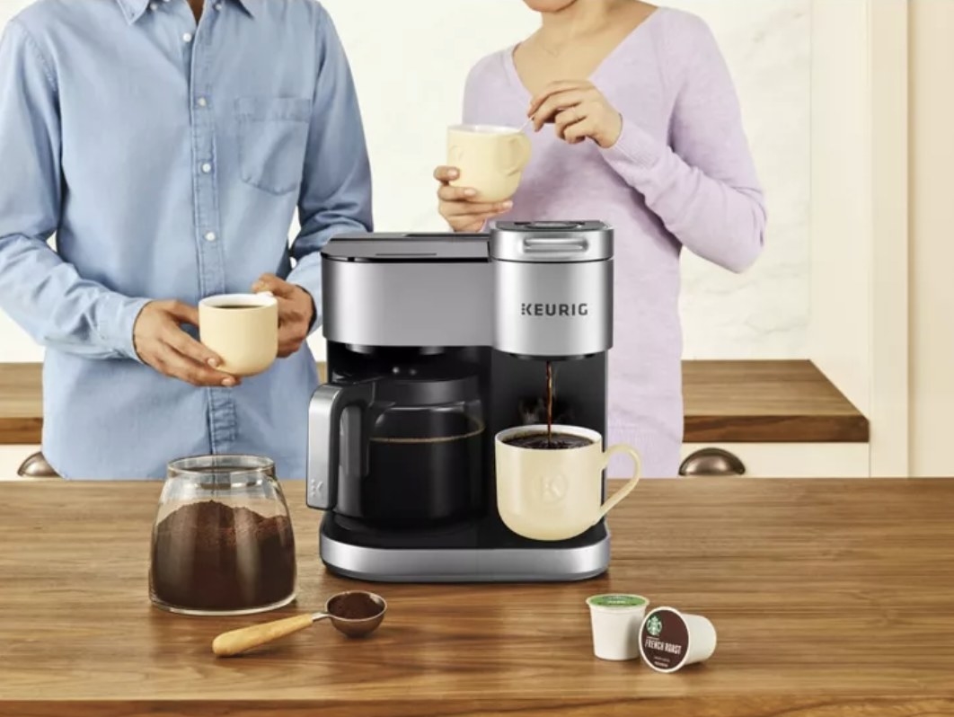 The Keurig machine is silver with black accents with two models are sipping their drinks in the background