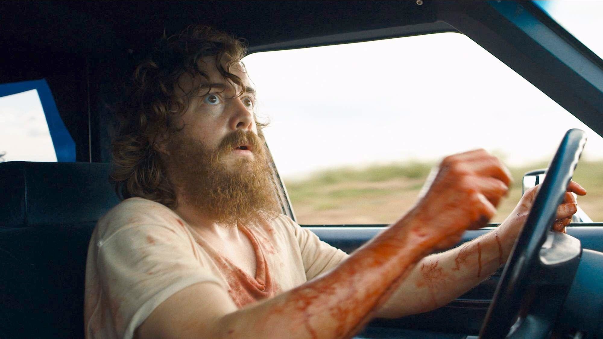 A bearded man covered in blood stares worriedly into his rearview mirror