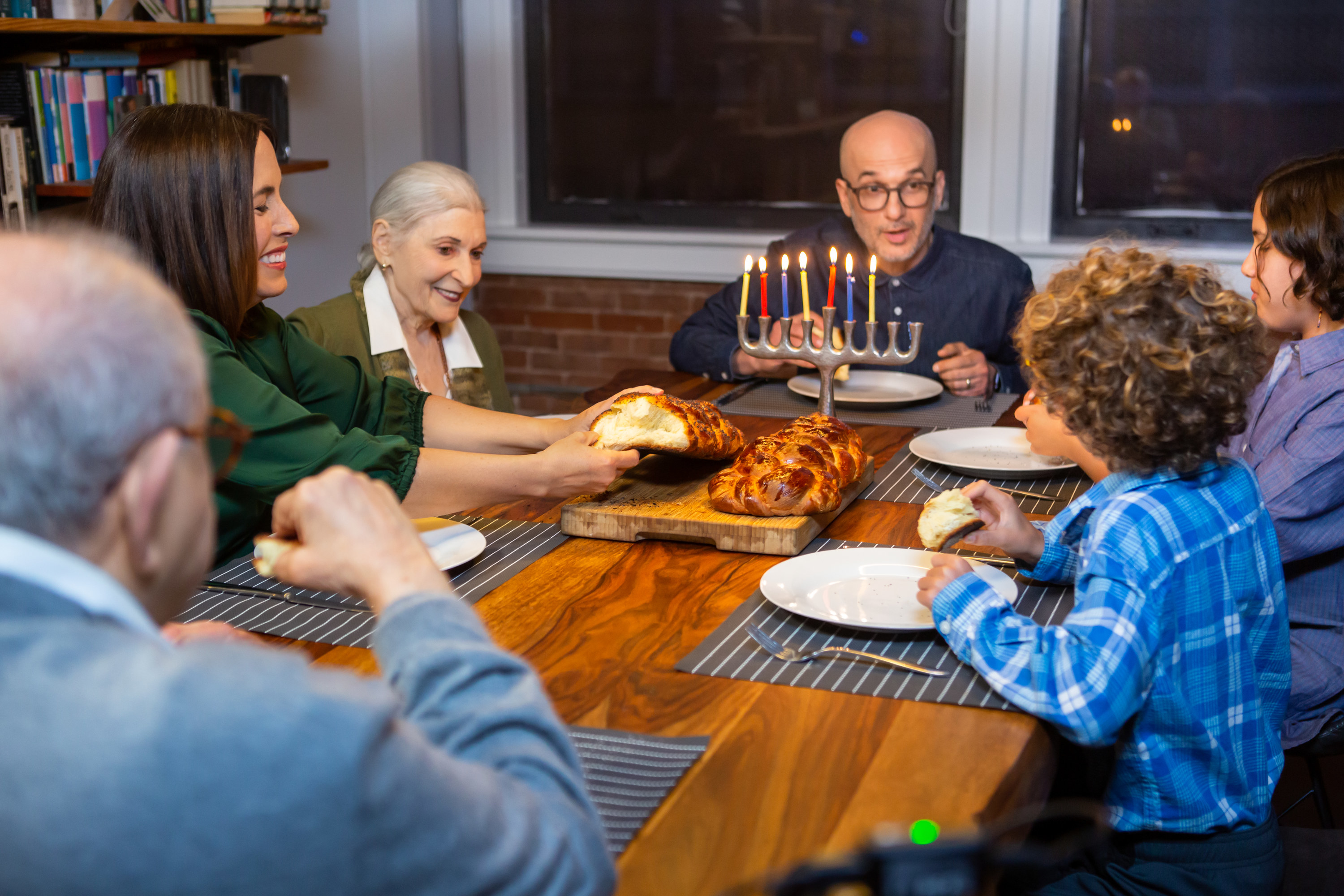 A mother tears challah while her family enjoys the Hanukkah festivities