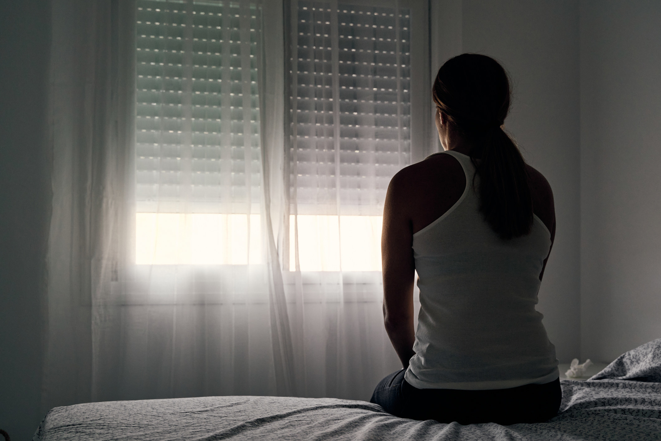 Woman sitting on a bed, staring out the window, and looking solemn