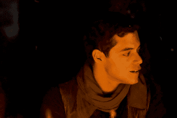 rami malek in an ochre-colored scarf staring into the fire